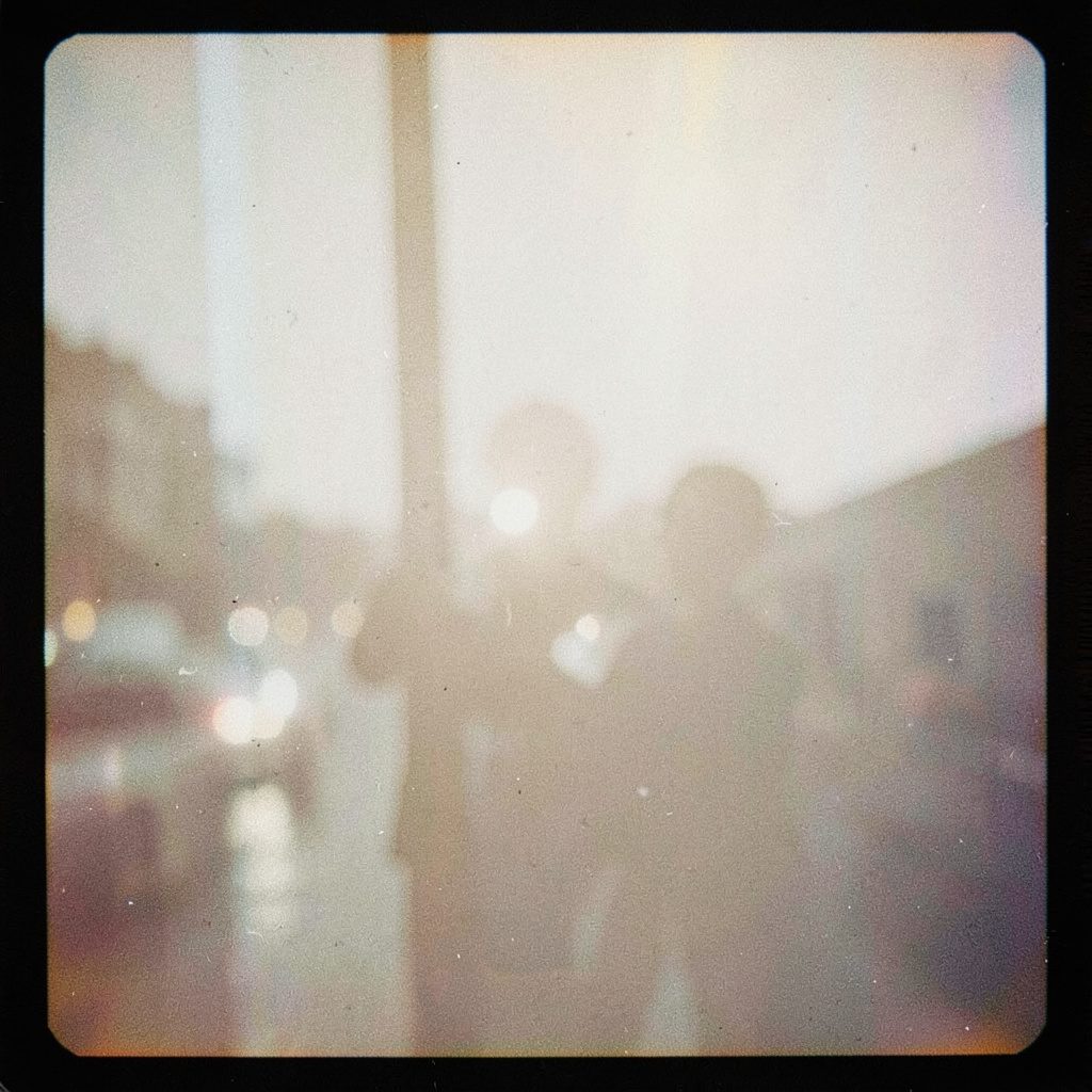 An analog film photograph with visible grain of a person smiling while holding a smartphone, warm amber and cream tones, cozy indoor setting