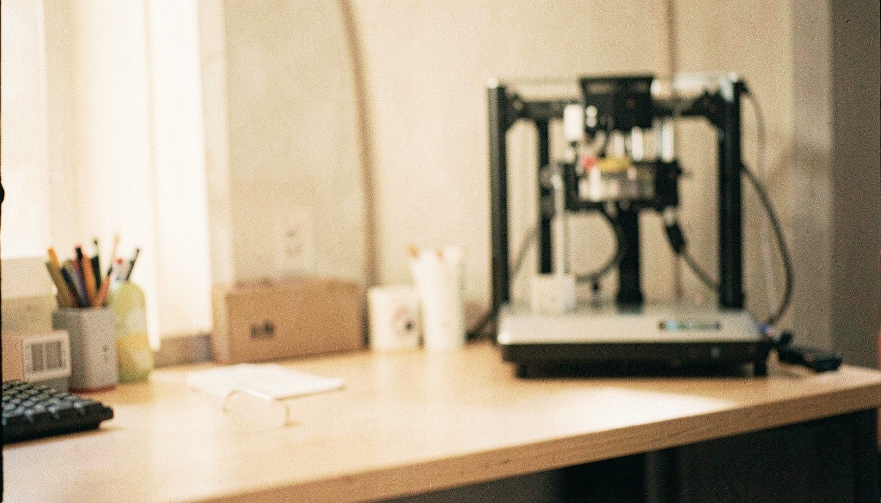 A warm-toned analog film photograph with visible grain showing a clean, modern desktop workspace with a 3D printer in soft focus in the background, warm amber and cream tones, calm and creative atmosphere.